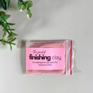 K&ouml;nnte beinhalten: Ein kleiner, durchsichtiger Plastikbeutel mit rosa Finish-Ton. Ein rosa Etikett auf dem Beutel tr&auml;gt die Aufschrift "The perfect finishing clay" mit zus&auml;tzlichem Text darunter. Der Beutel hat einen roten Streifen entlang des Rei&szlig;verschlusses.