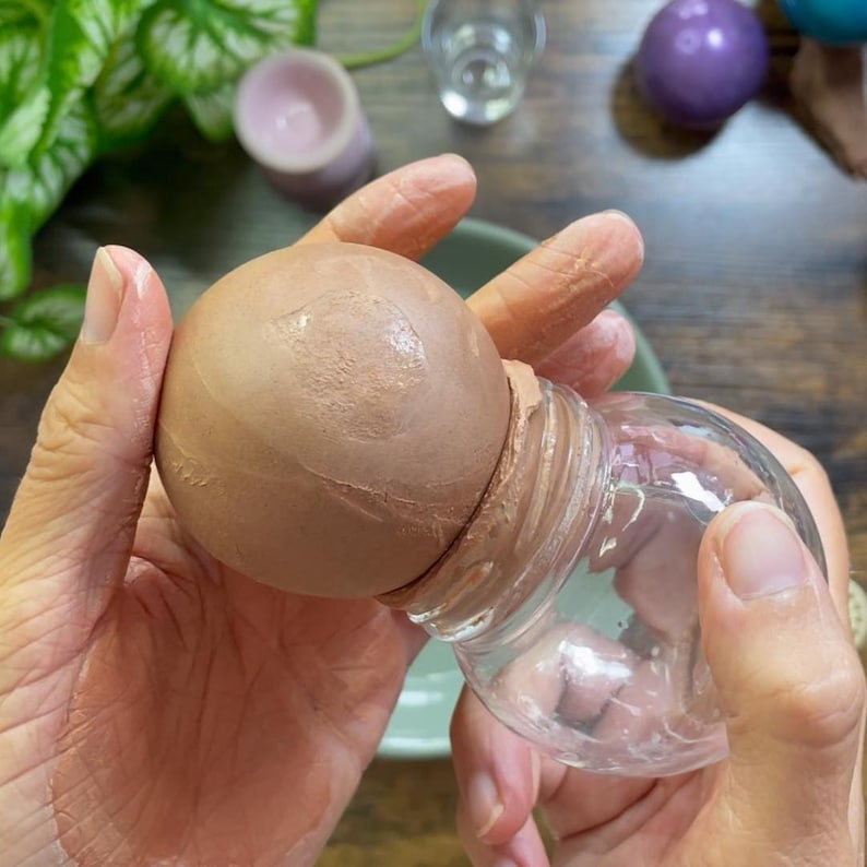 K&ouml;nnte beinhalten: Ein klares Glasgef&auml;&szlig; mit rundem Boden und einer braunen Kugel im Inneren. Die Kugel ist teilweise in das Gef&auml;&szlig; eingesetzt, wobei eine braune Substanz zwischen der Kugel und der &Ouml;ffnung des Gef&auml;&szlig;es sichtbar ist. Das Gef&auml;&szlig; wird von H&auml;nden gehalten.