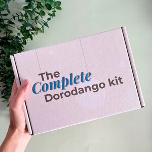 K&ouml;nnte beinhalten: Eine hellrosa rechteckige Box mit dem Text "The Complete Dorodango kit" in Blau und Dunkelgrau. Die Box wird von einer Hand gehalten, mit gr&uuml;nem Laub im Hintergrund.