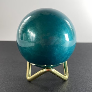 K&ouml;nnte beinhalten: Eine teal-farbene Kugel mit gl&auml;nzender Oberfl&auml;che, die auf einem kleinen, goldfarbenen Metallst&auml;nder ruht. Die Kugel reflektiert das Licht und erzeugt Glanzlichter. Der St&auml;nder hat ein einfaches, dreibeiniges Design. Die Gesamt&auml;sthetik ist modern und dekorativ.