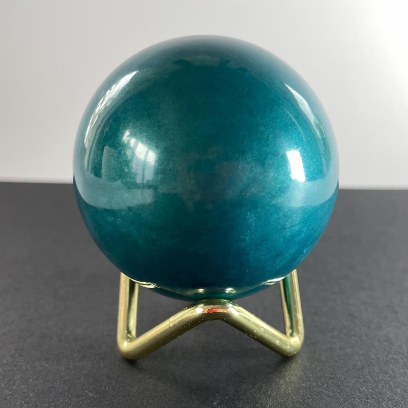 K&ouml;nnte beinhalten: Eine teal-farbene Kugel mit gl&auml;nzender Oberfl&auml;che, die auf einem kleinen, goldfarbenen Metallst&auml;nder ruht. Die Kugel reflektiert das Licht und erzeugt Glanzlichter. Der St&auml;nder hat ein einfaches, dreibeiniges Design. Die Gesamt&auml;sthetik ist modern und dekorativ.