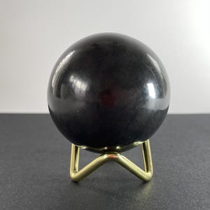 K&ouml;nnte beinhalten: Eine gl&auml;nzende schwarze Kugel, m&ouml;glicherweise ein dekorativer Stein, steht auf einem kleinen, goldfarbenen Metallst&auml;nder. Die Kugel reflektiert das Licht und erzeugt Glanzlichter. Der St&auml;nder hat ein dreibeiniges Design. Die Gesamt&auml;sthetik ist modern und minimalistisch.