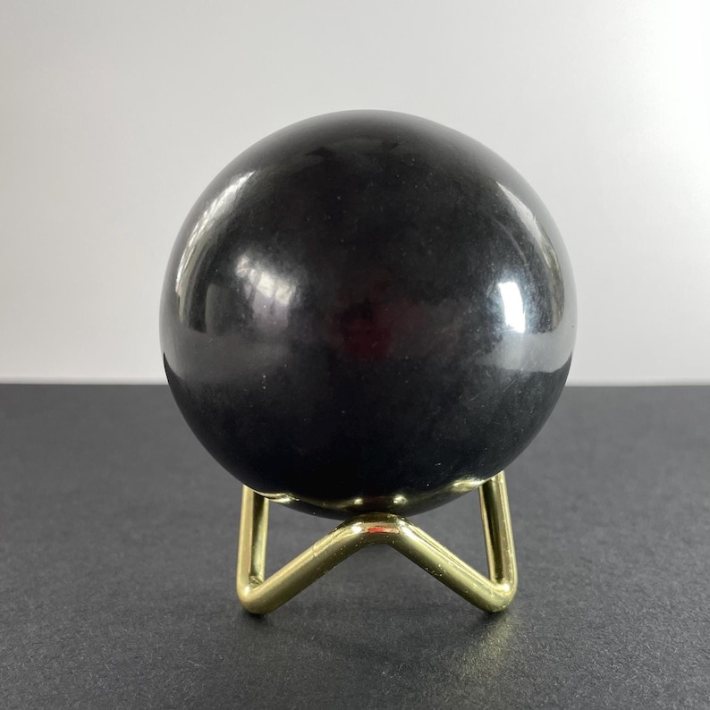 K&ouml;nnte beinhalten: Eine gl&auml;nzende schwarze Kugel, m&ouml;glicherweise ein dekorativer Stein, steht auf einem kleinen, goldfarbenen Metallst&auml;nder. Die Kugel reflektiert das Licht und erzeugt Glanzlichter. Der St&auml;nder hat ein dreibeiniges Design. Die Gesamt&auml;sthetik ist modern und minimalistisch.