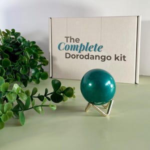 Può includere: Una sfera color verde acqua su un supporto dorato, con la scritta "The Complete Dorodango kit" su una scatola. Fogliame verde in primo piano. La sfera ha un diametro di circa 5 cm.