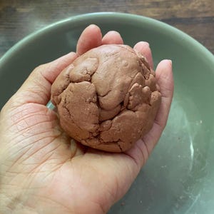 K&ouml;nnte beinhalten: Ein Ball aus r&ouml;tlich-braunem Teig, gehalten in einer Hand, mit einer hellgr&uuml;nen Sch&uuml;ssel im Hintergrund. Der Teig hat eine strukturierte, unebene Oberfl&auml;che. Das Bild ist gut beleuchtet und zeigt die Details des Teigs und der Sch&uuml;ssel.