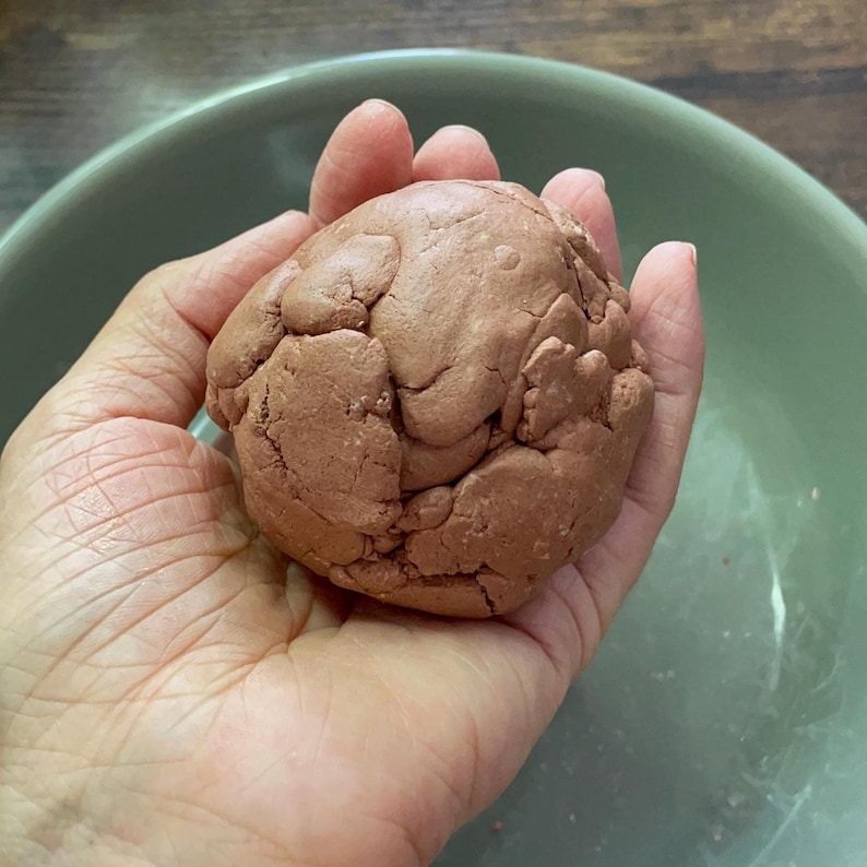 K&ouml;nnte beinhalten: Ein Ball aus r&ouml;tlich-braunem Teig, gehalten in einer Hand, mit einer hellgr&uuml;nen Sch&uuml;ssel im Hintergrund. Der Teig hat eine strukturierte, unebene Oberfl&auml;che. Das Bild ist gut beleuchtet und zeigt die Details des Teigs und der Sch&uuml;ssel.