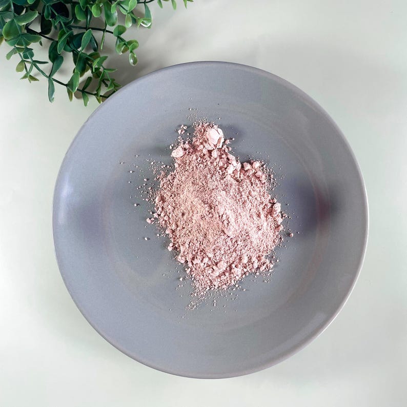 K&ouml;nnte beinhalten: Ein grauer Keramikteller mit einem Haufen rosa Pulver. Das Pulver hat eine helle, staubige Rosafarbe. Gr&uuml;ne Blattzweige befinden sich in der oberen linken Ecke des Bildes, und der Hintergrund ist wei&szlig;.