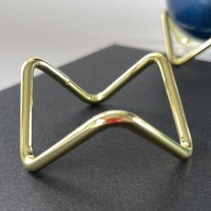 May include: Two gold-coloured metal stands with a geometric design. Each stand is made of bent metal rods, forming a triangular shape with a curved base. The stands are designed to hold a round object.