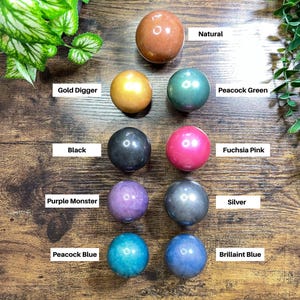 K&ouml;nnte beinhalten: Eine Auswahl an polierten Kugeln in verschiedenen Farben, darunter Braun, Gold, Gr&uuml;n, Schwarz, Pink, Lila, Silber, T&uuml;rkis und Blau. Jede Kugel hat eine gl&auml;nzende Oberfl&auml;che und ist mit Namen wie "Gold Digger" und "Brillaint Blue" beschriftet.