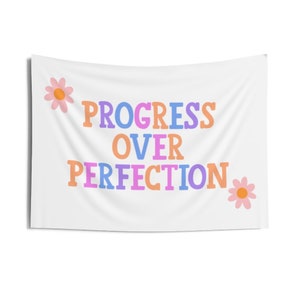 May include: White tapestry with the text "PROGRESS OVER PERFECTION" in colorful letters. Two pink flowers with yellow centers are on either side of the text.