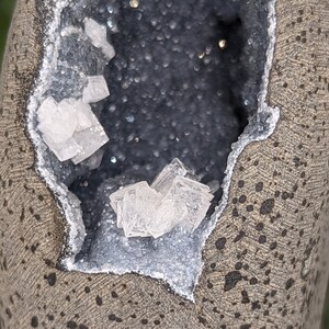 May include: A grey, round rock with a cavity revealing a cluster of white crystals on a black background.