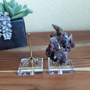 May include: Two clear acrylic display stands with a gold metal stand in each. One stand holds a large, iridescent, dark purple and brown crystal cluster. The stands have a triangular logo on the bottom.
