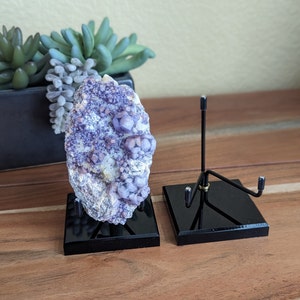 May include: A large, rough-hewn mineral specimen with a cluster of small, spherical, purple crystals. The mineral sits on a black display stand. A second stand is next to it. A succulent plant is in the background.