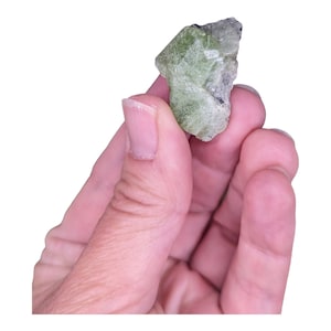 May include: A green, rough, and irregularly shaped crystal with a dark green color. The crystal is being held in a hand.