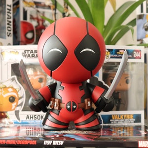 Peut inclure: Une figurine Funko Pop! de Deadpool rouge et noire avec un grand sourire, tenant deux épées argentées. La figurine est posée sur une bande dessinée avec les mots "Spider-Man/Deadpool Itsy Bitsy" imprimés sur la couverture.