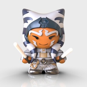 May include: A small, orange and white figurine of a Star Wars character with blue and white headwear, holding two white lightsabers.