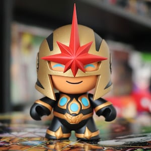 Op de afbeelding: Een zwart en gouden action figure met een rode ster op zijn helm. De figuur heeft een blauw en gouden borststuk en staat op een gepatroneerd oppervlak.