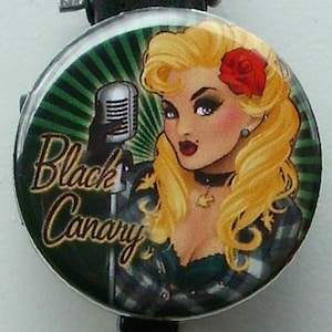 May include: A black and green retractable badge holder with a round, colorful button featuring a cartoon woman with blonde hair, a red flower, and a microphone. The text "Black Canary" is written on the button.