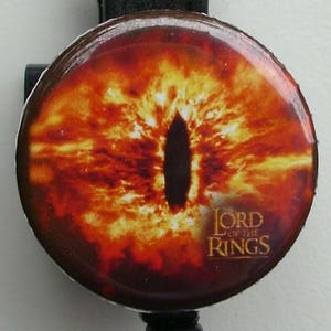 Könnte beinhalten: Ein runder Knopf mit einem feurigen orangefarbenen und gelben Auge mit einer schwarzen Pupille. Der Text "Lord of the Rings" ist in Gold am unteren Rand des Knopfes aufgedruckt.