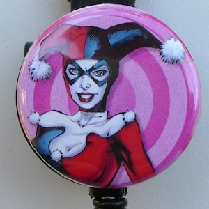 May include: A pink and white circular badge with a graphic of Harley Quinn, a comic book character, wearing a red and black jester costume. The badge is attached to a black retractable badge reel.
