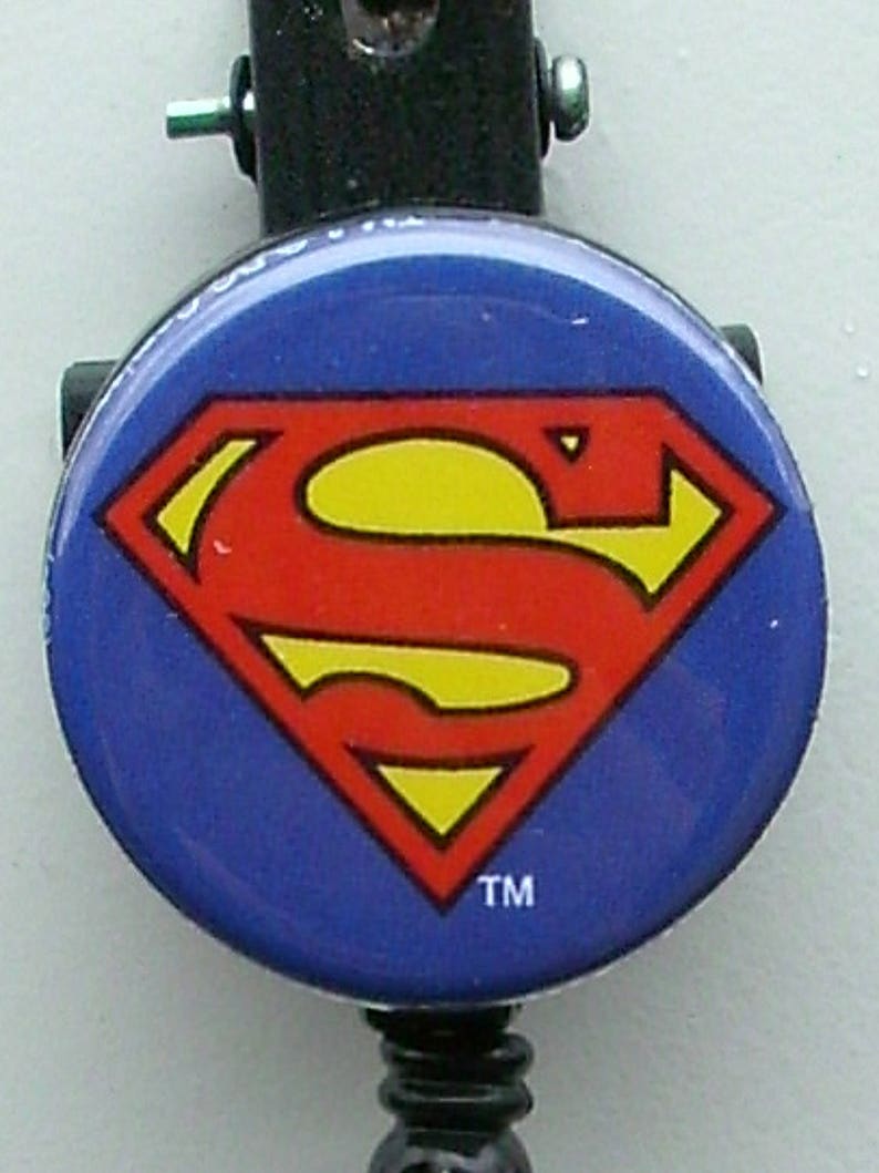 Puede incluir: Una insignia redonda con el logotipo de Superman, un diamante rojo y una 'S' amarilla sobre fondo azul. Sujeta a un carrete retr&aacute;ctil negro con clip.
