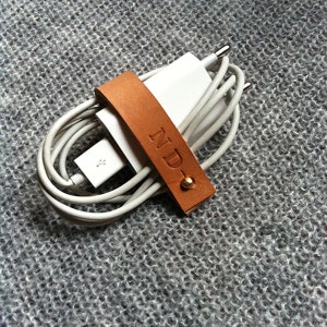 Peut inclure: Un organisateur de cordon en cuir marron avec les initiales "ND" gravées dessus. L'organisateur contient un chargeur de téléphone blanc et un cordon.