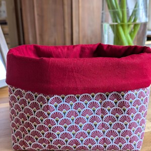 Peut inclure: Un panier rectangulaire en tissu avec un intérieur rouge et un extérieur à motifs. L'extérieur présente un motif répétitif en forme d'éventail en rouge, blanc et or. Le panier est posé sur une surface en bois.
