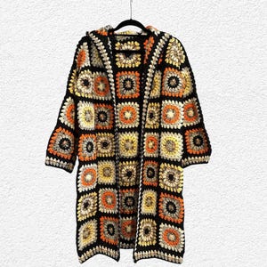 May include: A long, open-front hooded cardigan in a granny square pattern. The crochet jacket features a black base with squares of orange, yellow, and cream. The sleeves are three-quarter length, and the garment hangs from a black hanger.