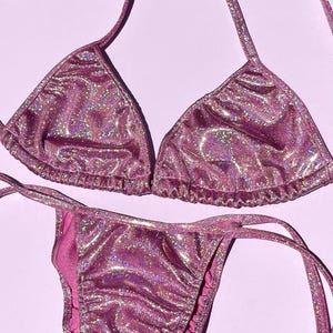 May include: A sparkly pink bikini set featuring a triangle top and string tie bottoms. The material has a glittery, iridescent finish. The swimwear is presented on a light purple backdrop.