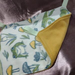 May include: A small, square baby blanket with a white background, featuring a pattern of green frogs, yellow mushrooms, and blue flowers. The blanket has a yellow fleece backing and light green ribbon ties.