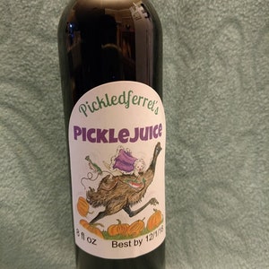 Puede incluir: Una botella de vidrio oscuro con una etiqueta blanca que dice "Pickledferret's PickleJuice". La etiqueta presenta una ilustraci&oacute;n de dibujos animados de un em&uacute; con una capa morada y llevando un peque&ntilde;o animal blanco. La botella es de 8 fl oz y tiene una fecha de caducidad del 12/1/18.