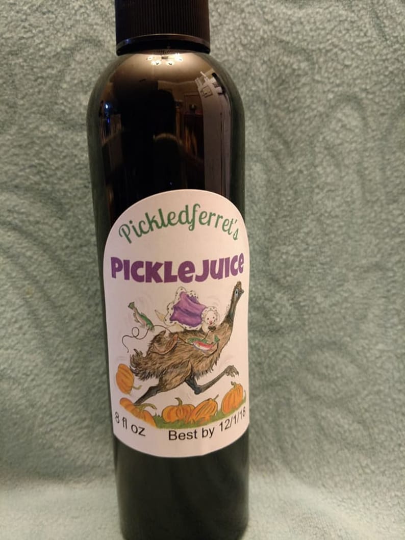 Puede incluir: Una botella de vidrio oscuro con una etiqueta blanca que dice "Pickledferret's PickleJuice". La etiqueta presenta una ilustraci&oacute;n de dibujos animados de un em&uacute; con una capa morada y llevando un peque&ntilde;o animal blanco. La botella es de 8 fl oz y tiene una fecha de caducidad del 12/1/18.