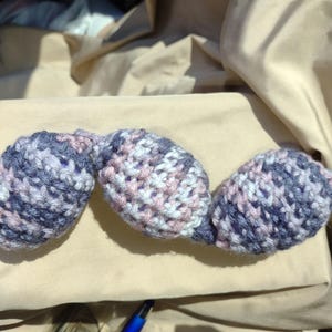 May include: A handmade, crocheted item in shades of blue, white, and pink. The item appears to be three connected oval shapes, possibly a decorative piece or part of a larger craft project. The yarn has a textured appearance.