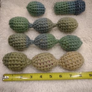 May include: Crocheted items in shades of green and blue, including a small ball, a cylinder, and three sets of connected spheres. A yellow tape measure shows the items' size. The items are handmade.