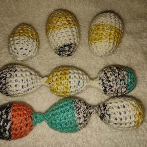 May include: A collection of handmade crochet toys in various colors. The toys include egg-shaped and dumbbell-shaped designs. Colors include white, yellow, black, orange, and teal. The toys are arranged on a soft, textured surface.
