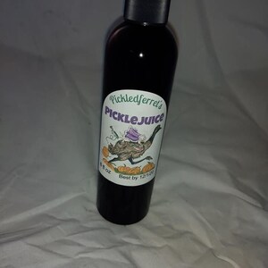 Puede incluir: Una botella negra con una etiqueta que dice "Pickledferret's Picklejuice". La etiqueta presenta un em&uacute; de dibujos animados con un sombrero morado y llevando una peque&ntilde;a bolsa morada. La botella es de 8 fl oz y tiene una fecha de caducidad del 12/1/2020.