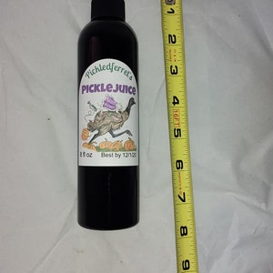Puede incluir: Una botella de vidrio negro con una etiqueta blanca que dice "Pickledferret's PICKLE JUICE" y presenta una ilustraci&oacute;n de dibujos animados de un em&uacute; con un vestido morado y montando una calabaza. La etiqueta tambi&eacute;n dice "8 fl oz Best by 12/1/20".