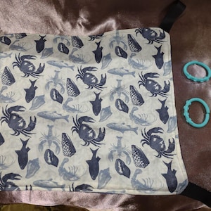 May include: A white and blue patterned fabric with crabs, fish, and other sea creatures. The fabric is attached to black straps. Four light blue teething rings are also visible.