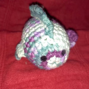 May include: A crocheted stuffed fish toy with a purple, white, and teal color scheme. The fish has a small black eye and a fin on its back.