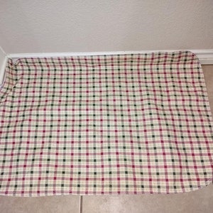 May include: A cream-colored checkered rug with red and green squares. The rug has a frayed edge and is made of a soft, woven fabric.