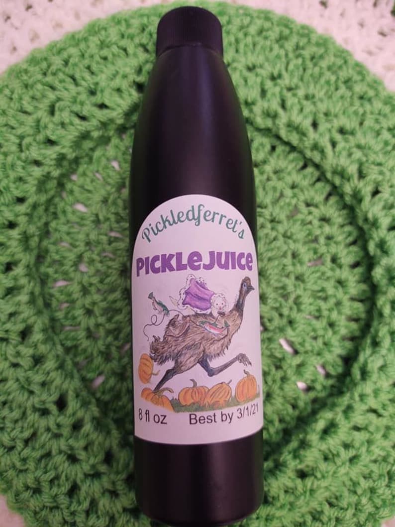 Puede incluir: Una botella negra con una etiqueta blanca que dice "Pickledferret's PickleJuice". La etiqueta tiene una ilustraci&oacute;n de dibujos animados de un em&uacute; con un vestido morado y llevando peces. La botella es de 8 fl oz y tiene una fecha de caducidad del 3/1/21.