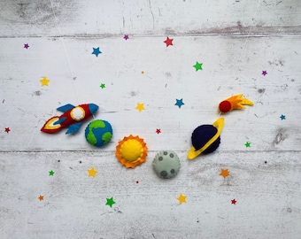 May include: A felt rocket, planets, and a comet mobile hanging from a white string against a white wood background. The mobile features a red and blue rocket, a blue and green planet, a yellow sun, a gray moon, and a blue planet with a yellow ring. The background is decorated with colorful felt stars.