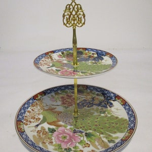 May include: Two-tiered serving stand with a gold metal base and a decorative top. The stand features two round porcelain plates with a peacock and floral design in shades of blue, green, gold, and pink.
