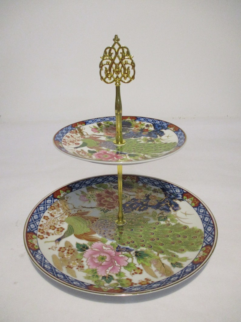 May include: Two-tiered serving stand with a gold metal base and a decorative top. The stand features two round porcelain plates with a peacock and floral design in shades of blue, green, gold, and pink.