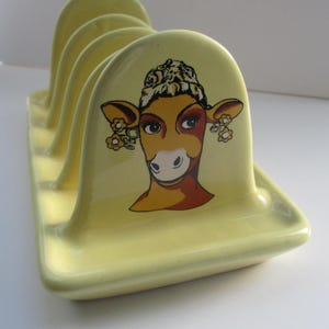 May include: A yellow ceramic toast rack featuring a cartoon cow face design. The cow has a floral headband and earrings.