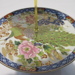 May include: A white ceramic plate with a blue and gold border. The plate features a colorful design of peacocks and flowers.