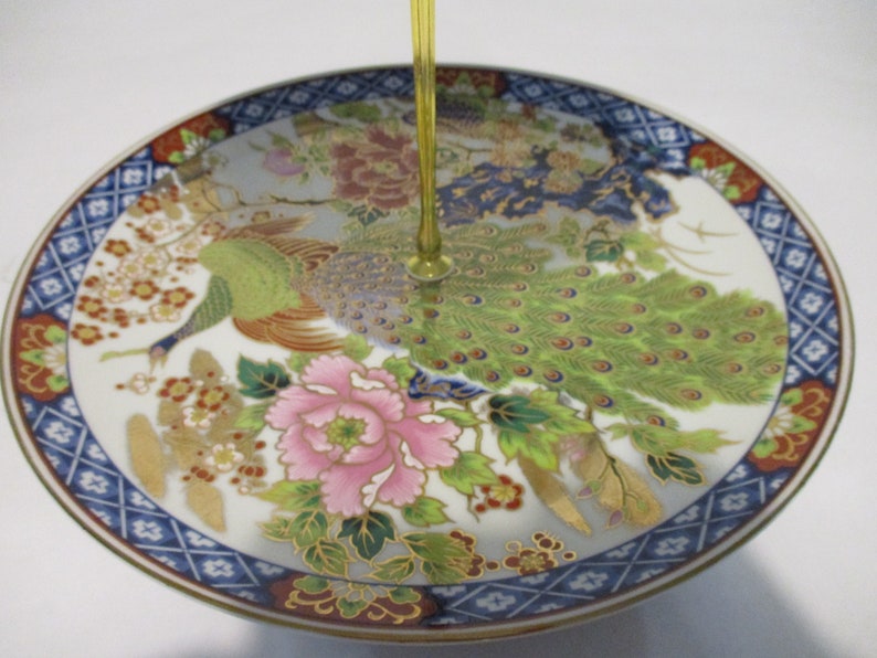 May include: A white ceramic plate with a blue and gold border. The plate features a colorful design of peacocks and flowers.
