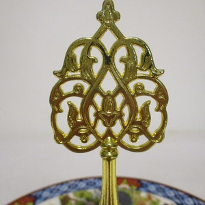 May include: A gold metal decorative stand with a round top and three legs. The top of the stand features a decorative design with intricate details.