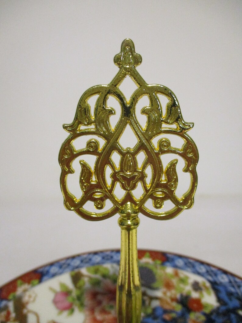 May include: A gold metal decorative stand with a round top and three legs. The top of the stand features a decorative design with intricate details.