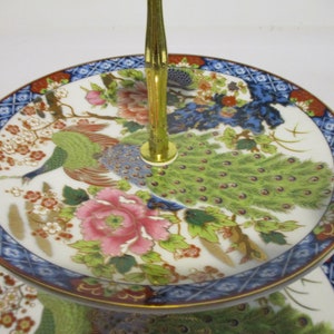 May include: Two-tiered porcelain serving stand with a gold metal handle. The stand features a white background with a colorful peacock design and floral accents.